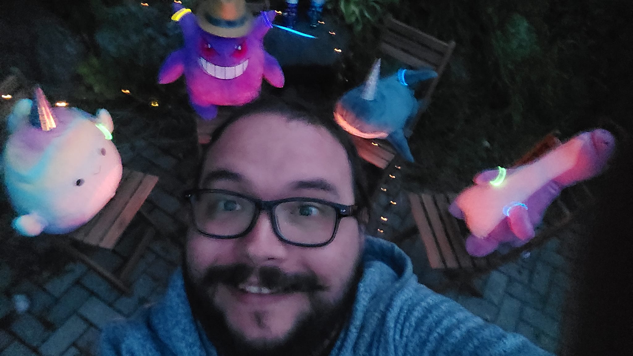 A man with facial hair stands in front of a stuffed animal campfire, the photo is taken from above and his head blocks the fire, four stuffed animals are visible